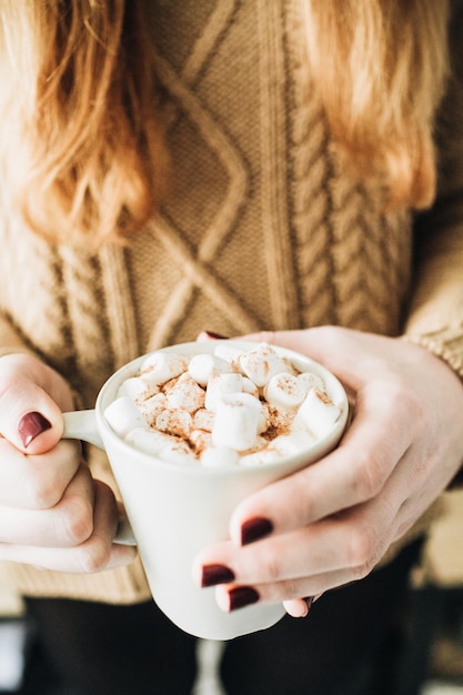 Tasse avec café chaud avec crème fouettée guimauve et cannelle dans les mains des femmes Belle femme aux cheveux rouges tenir dans ses mains mug