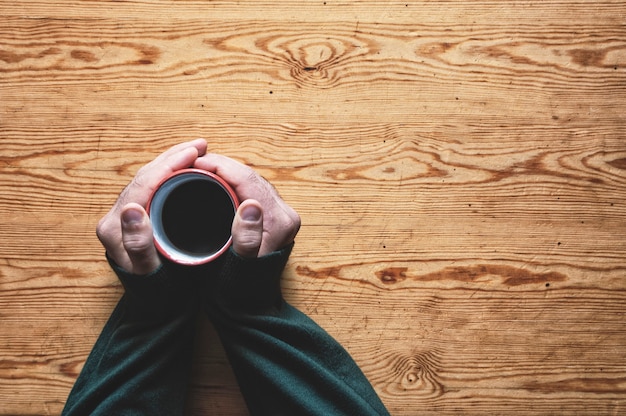 Une tasse de café chaud entre les mains d'un homme sur une surface en bois à gauche.