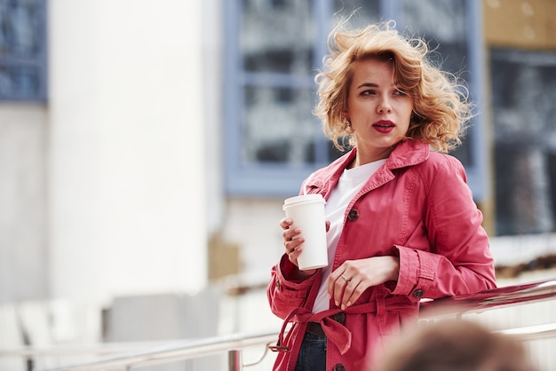 Avec une tasse de café chaud. Jolie femme adulte en manteau rouge chaud se promène dans la ville à son heure du week-end