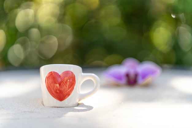 tasse de café avec coeur rouge imprimé sur du sable sur fond de fleur flou.