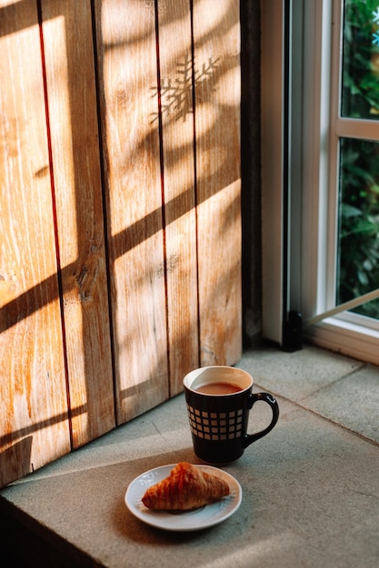 Tasse de café confortable et un croissant sur fond de fenêtre de jour ensoleillé avec des ombres de soleil