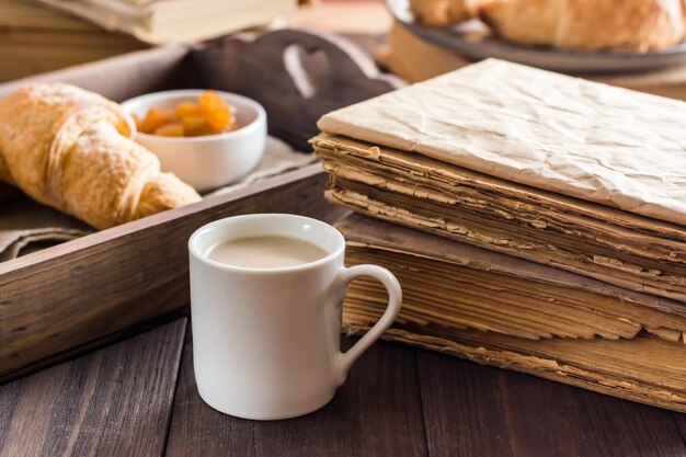 Une tasse de café à côté d'une pile de vieux livres et d'un plateau avec des croissants sur une table en bois. Détente, bien-être et lecture à domicile.