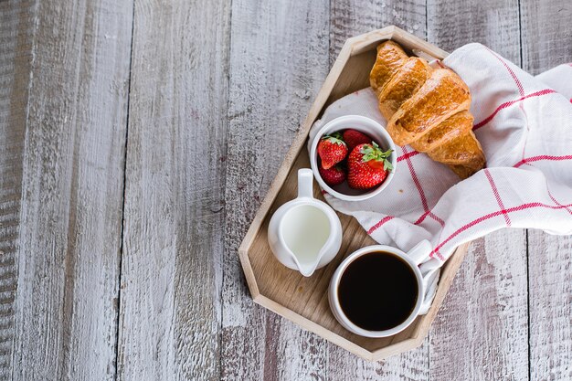 Tasse de café, croissants frais et fraises fraîches sur un plateau en bois.