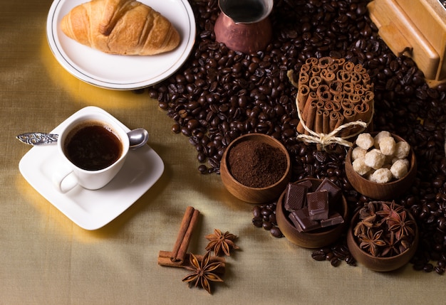 Tasse à café, cruissant, grains de café, cannelle, chocolat et sucre en cubes sur une table