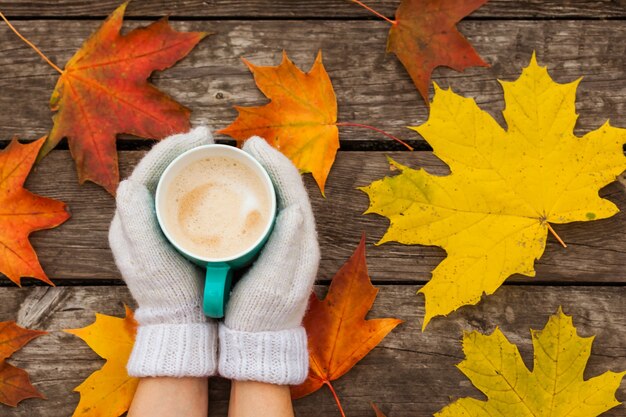 La tasse de café dans les mains gelées. à la surface des feuilles d'automne jaunes. Sur une surface en bois sombre.
