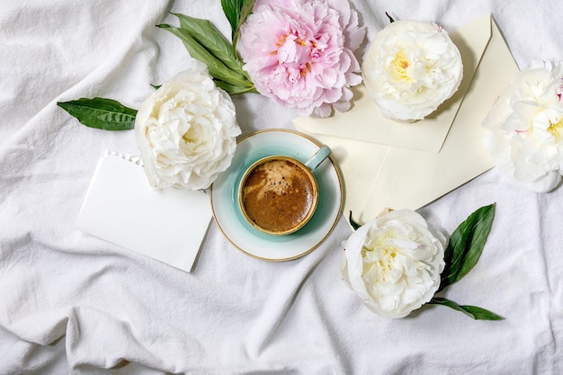 Tasse de café expresso, papier vierge, enveloppe, fleurs de pivoines roses et blanches avec des feuilles sur une surface textile en coton blanc