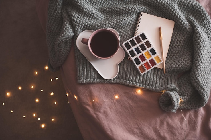 Photo tasse de café frais sur une assiette avec palette de peintures, carnet de croquis et pull tricoté sur des lumières de noël rougeoyantes à l'arrière-plan en gros plan sur le lit. l'hiver.