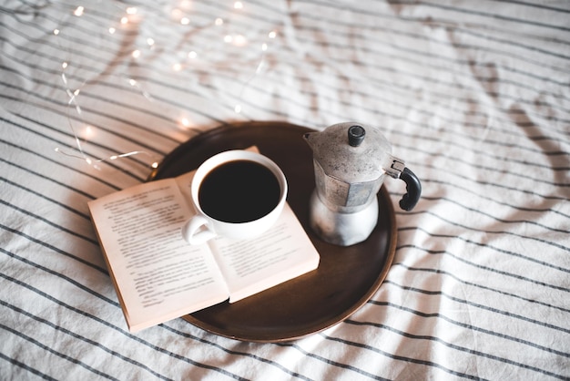 Tasse de café frais sur un livre ouvert avec une cafetière restant sur un plateau en bois en gros plan. Bonjour.