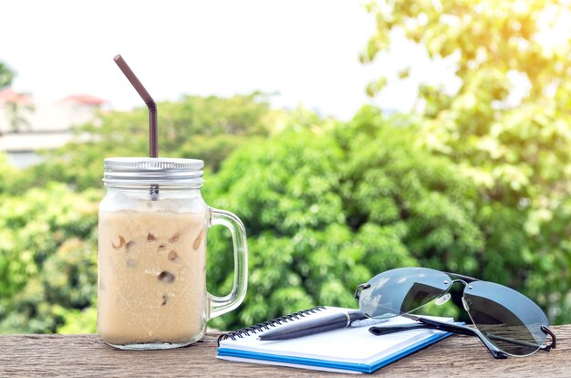 Tasse à café de glace avec poignée avec un cahier, des lunettes de soleil et un stylo sur l'ancien plancher en bois.