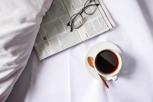 Une tasse de café et un journal sur un lit blanc le matin.