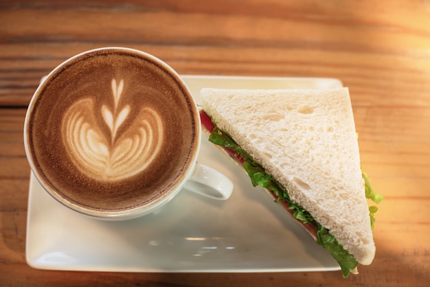 Une tasse de café avec motif coeur dans une tasse blanche et un sandwich sur fond de table en bois