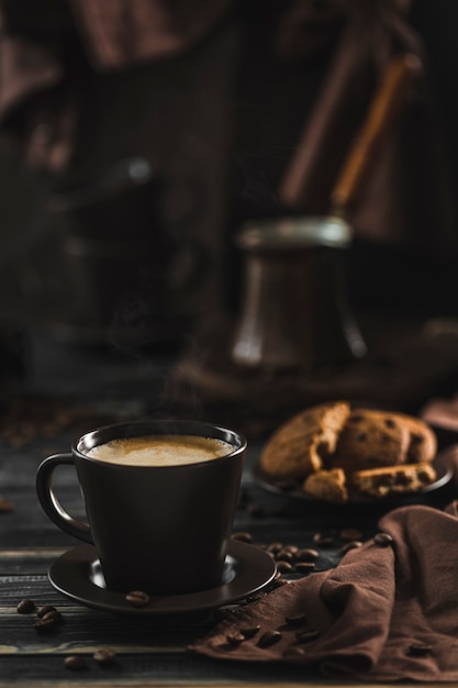 Une tasse de café avec de la mousse sur un bois sombre avec des biscuits à l'avoine, des grains épars, une cafetière