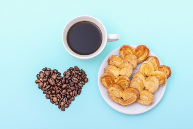 Tasse de café noir, biscuits en forme de coeur, grains de café en forme de coeur sur fond bleu