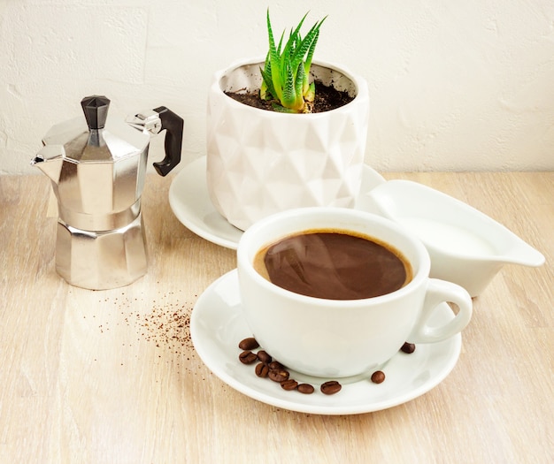 Tasse de café noir de couleur blanche avec soucoupe et haricots, cafetière en aluminium, récipient à lait avec du lait, plante succulente verte en pot blanc sur table en bois beige. Mise au point sélective. Copier l'espace