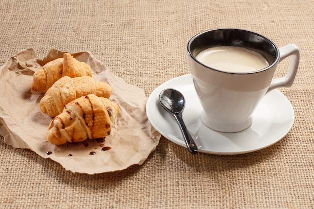 Tasse de café noir avec cuillère sur soucoupe, croissants frais au chocolat sur papier d'emballage avec un sac sur le fond.