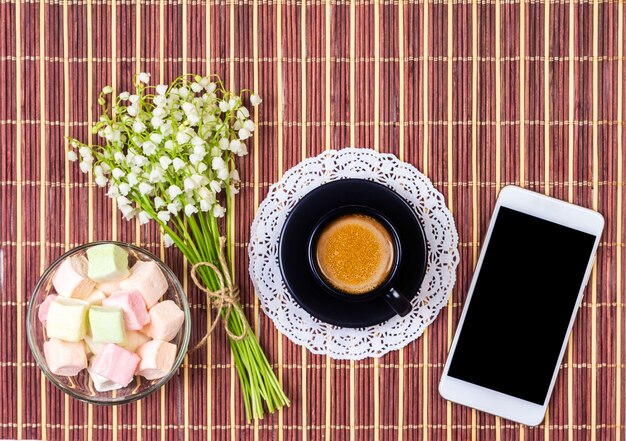 Tasse de café noir avec du lait sur une soucoupe, guimauve, un bouquet de fleurs de muguet et un smartphone sur la table, vue de dessus à plat