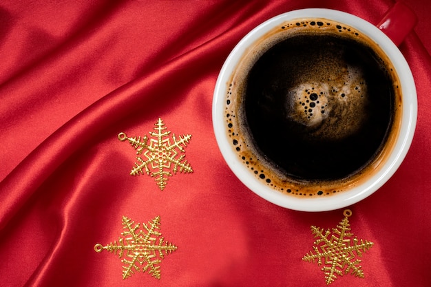 Une tasse de café noir avec des ornements d'étoiles de Noël sur un fond de tissu de soie rouge vif.