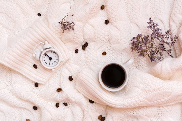 Une tasse de café noir, un réveil, des fleurs séchées et des grains de café sur un confortable pull blanc. Concept de temps de bien-être. Vue de dessus