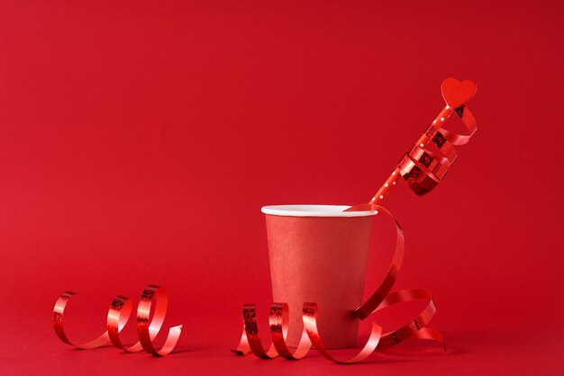 Tasse à café en papier avec coeur en bois sur paille et rubans festifs sur surface rouge. Concept de la Saint-Valentin