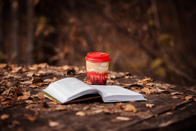 une tasse de café en papier et un livre ouvert sur une table en bois parmi les feuilles tombées