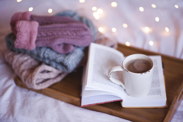 Tasse de café restant sur un livre ouvert avec une pile de vêtements tricotés gants bonnet écharpe sur un plateau en bois au-dessus des lumières à l'arrière-plan Saison d'hiver Ambiance chaleureuse