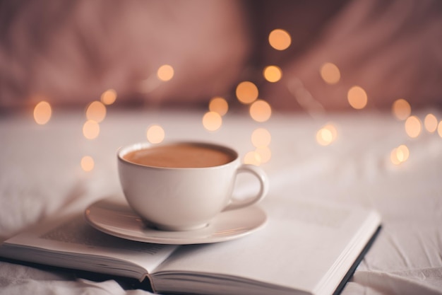 Tasse de café restant sur un livre de pepaer ouvert sur des lumières de Noël rougeoyantes se bouchent. Bonjour. L'heure du déjeuner. Noël. Temps de vacances d'hiver.