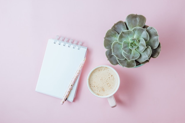 Tasse à café rose du matin, cahier propre, stylo et fleur en pot sur fond rose