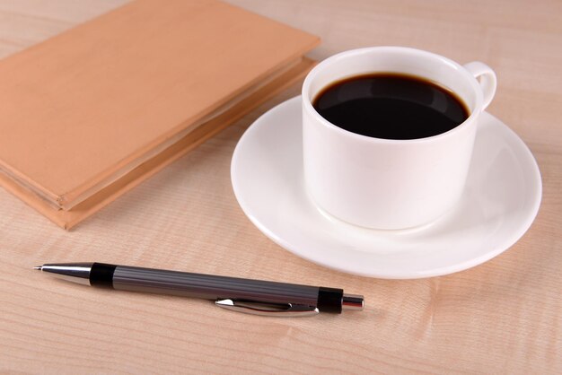 Tasse de café sur une soucoupe avec un cahier fermé et un stylo sur fond de table en bois