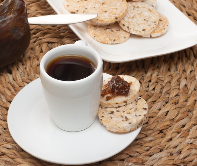 Une tasse de café sous la table de paille entourée d'un bol rempli de biscuits au riz et d'un pot de gelée