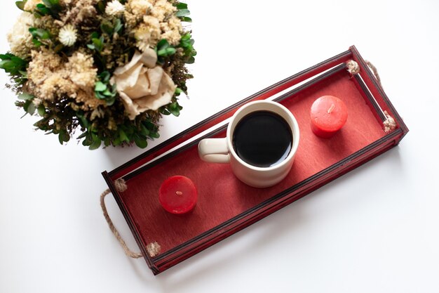 Une tasse de café ou de thé, des fleurs séchées et des bougies blanches et rouges, une boîte en bois rouge sur la fenêtre.