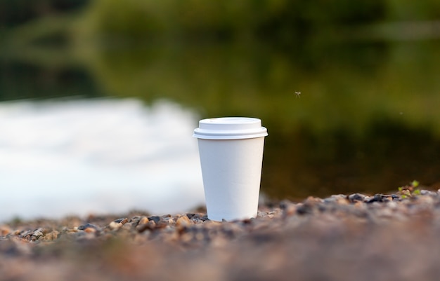 Une tasse de café ou de thé en papier blanc se dresse sur la rive rocheuse du lac. Une tasse de boisson chaude sur un caillou. Fond de belle nature du lac dans la forêt derrière. espace de copie gratuit.
