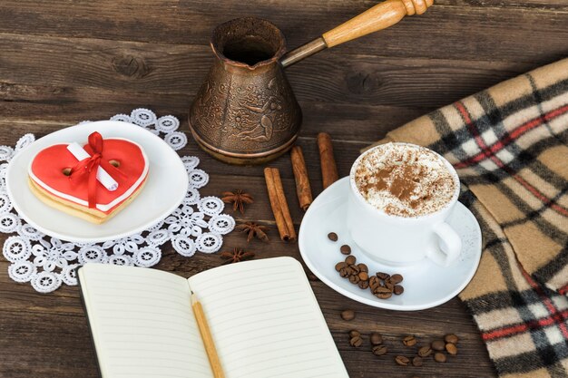 Tasse de cappuccino, message de largeur de biscuits en forme de coeur, cahier, crayon et cafetières sur une table en bois marron. Planification des vacances