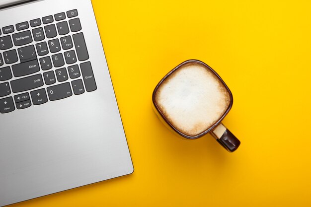 Tasse de cappuccino et ordinateur portable sur fond jaune. Concept indépendant, espace de travail, travail en ligne. Vue de dessus