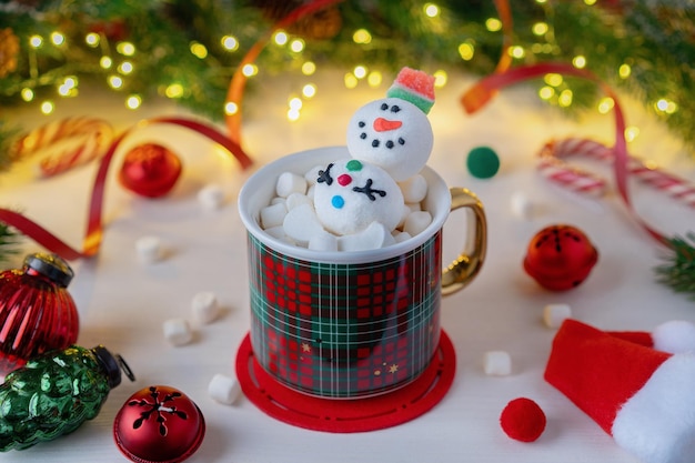 Une tasse à carreaux rouge avec bonhomme de neige et guimauves sur fond de décorations de Noël et de branches de sapin