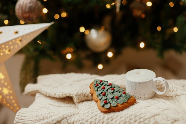 Tasse chaude de café et biscuit de pain d'épice de sapin de Noël sur un pull tricoté sur fond de sapin de Noël avec des lumières Bannière de Noël hygge d'hiver atmosphérique Vacances confortables