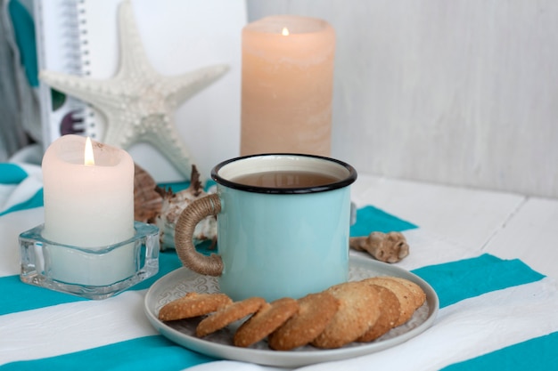 tasse chaude de thé noir avec des biscuits maison sur une nappe à rayures, des bougies de cire, des étoiles de mer décoratives, des coquillages, des tas de cahiers
