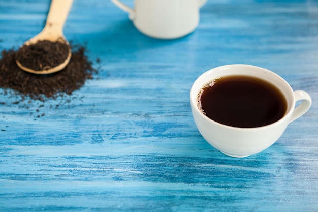 Tasse avec du thé chaud à côté d'une cuillère en bois avec des feuilles de thé sur fond bleu vintage