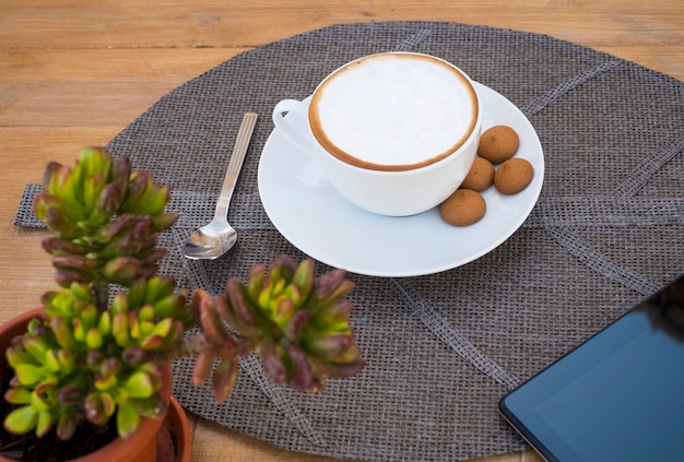 Tasse invitante de cappuccino avec mousse de lait et croissant frais sur un napperon en paille sur une table en bois - succulente et tablette dans les coins