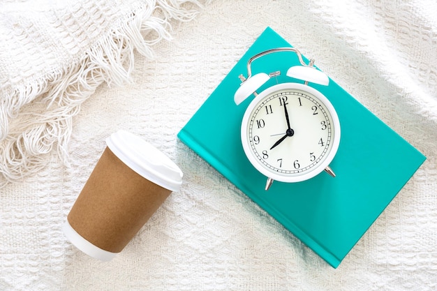 Tasse jetable en papier réveil blanc et livre vert en vue de dessus de lit blanc