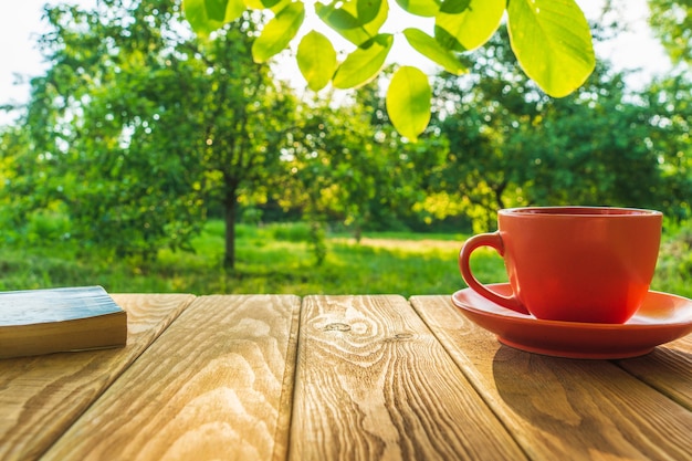 Tasse orange de café chaud et un livre sur une table en bois dans un jardin verdoyant le matin