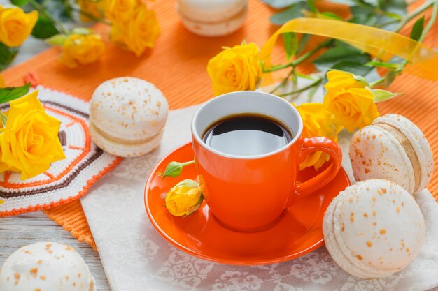 Tasse orange de café noir, fleurs de roses jaunes et macarons français pastel sucrés sur table en bois blanc