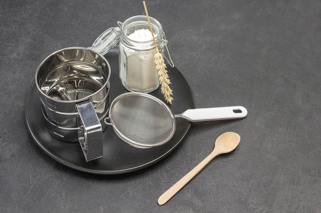 Tasse pour tamiser la farine et petit pot en verre à tamis avec de la farine sur plaque noire Spikelets de blé