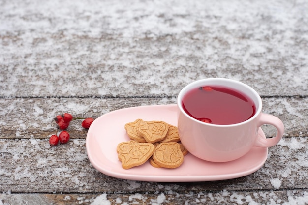 Une tasse rose de thé à l'églantier se dresse sur une surface en bois en hiver, le gel est visible