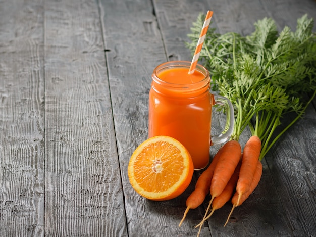 Une tasse de smoothie aux carottes fraîches avec de la paille cocktail, des carottes et des oranges sur une table rustique.