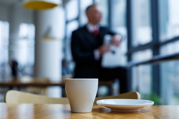 Une tasse et une soucoupe blanches avec du café aromatique chaud se dressent sur une table en bois dans le contexte d'un homme d'affaires assis