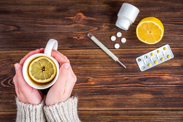 Tasse de thé au citron entre les mains des femmes. Citron, thermomètre et pilules sur une surface en bois. Traitement au froid. Saison de la grippe.