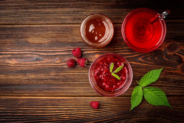 Tasse de thé aux framboises avec confiture de framboises et miel sur fond de bois foncé.