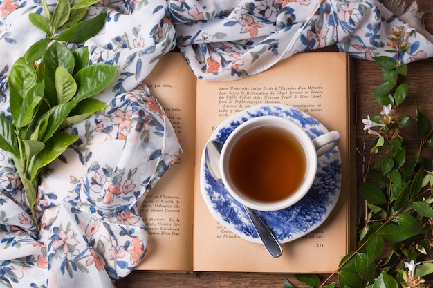 Photo tasse à thé aux herbes et soucoupe sur un livre ouvert avec feuilles et écharpe sur la table