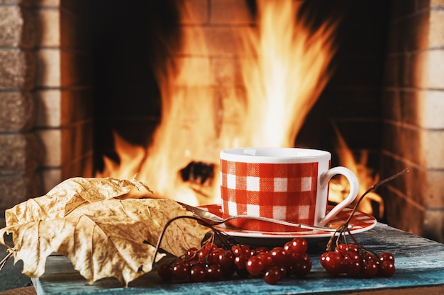 Une tasse de thé ou de café, des feuilles d'automne et des baies de viorne près d'une cheminée chaleureuse, dans une maison de campagne, des vacances d'automne ou d'hiver.