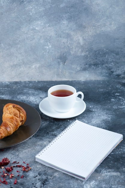 Une tasse de thé chaud avec un cahier ouvert et un croissant frais.